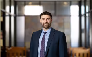 A smiling portrait of a bearded man with a dark blue suit and purple tie. 