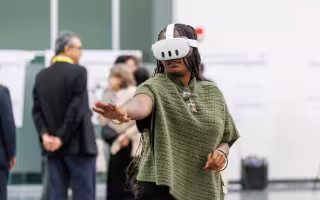 A woman with long braids and a green sweater wears a virtual reality headset and gestures in front of her.