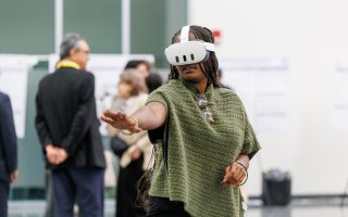 A woman with long braids and a green sweater wears a virtual reality headset and gestures in front of her.