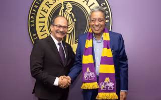 A smiling man in a blue blazer, gold and purple UAlbany scarf shakes hands with a smiling man in a black suit and purple tie.