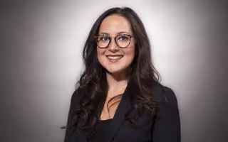 A woman with long dark hair and glasses in a black blazer smiles for a portrait against a gray backdrop.