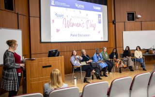 Seven women are seated on a stage as another woman asks questions from a lectern. A screen behind them says "International Women's Day Panel Discussion.""