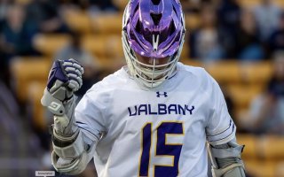 A man wears a UAlbany white lacrosse uniform with the number 15 and lacrosse gloves and a purple helmet.