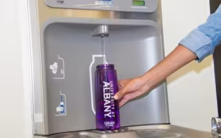 An arm and hand are seen refilling a water bottle with the words University at Albany printed on it