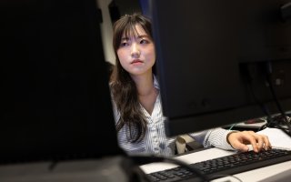 A young woman with dark hair wearing a collared button-down blue and white shirt looks at a pair of monitors in the UAlbany AI for Business Lab