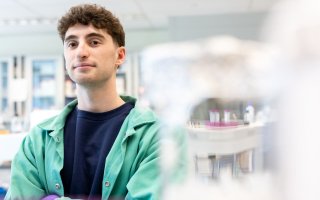 A young man with dark hair wearing a green lab coat stands in a brightly lit lab. The background is artfully blurred. 