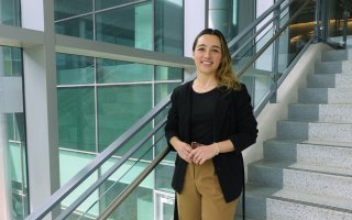 A young woman with brown-blond ombre hair, wearing a black top, black blazer and khaki slacks, smiles for a portrait in a stairwell with glass walls and gentle natural light.