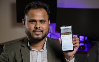 A man in a gray suit jacket holds up a phone with an app on the screen