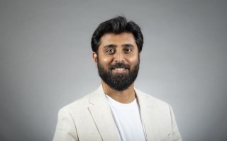 A man with dark brown hair wearing a white T-shirt with white linen blazer smiles for a portrait against a gray backdrop.