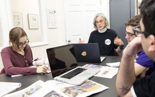 A group of students sit at a table with laptops discussing journalism and articles with a professor wearing a blue sweater.
