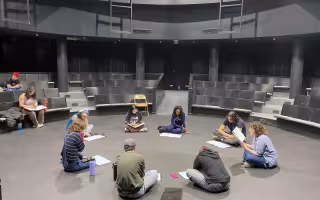 A group of students sit in a theatre-in-the-round with dark seats and walls as they sit in a circle with scripts in front of them.