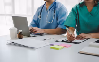 Two people wearing scrubs sit at a white desk. One is working on a laptop, wearing a stethoscope. The other is writing notes on a clipboard. Papers, post-it pads, and several medicine bottles are also on the table.