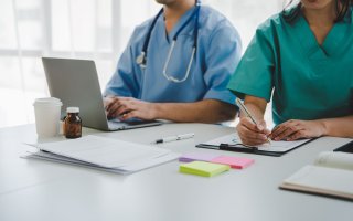 Two people wearing scrubs sit at a white desk. One is working on a laptop, wearing a stethoscope. The other is writing notes on a clipboard. Papers, post-it pads, and several medicine bottles are also on the table.