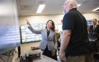 A woman with brown, shoulder-length brown hair and wearing a gray blazer points to a wall-mounted computer screen while a man in a black polo shirt looks on from the foreground.