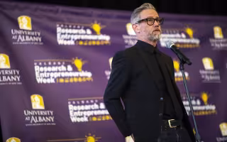 A bearded man with gray/black hair, dark-rimmed glasses and dressed all in black stands at a microphone in front of a purple background that includes the words "University at Albany" and "Research & Entrepreneurship Week"
