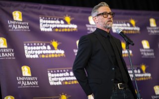A bearded man with gray/black hair, dark-rimmed glasses and dressed all in black stands at a microphone in front of a purple background that includes the words "University at Albany" and "Research & Entrepreneurship Week"