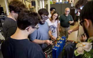 Around half a dozen people in safety glasses gather in a laboratory around a battery as a man holds various wires.