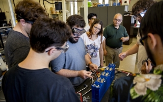 Around half a dozen people in safety glasses gather in a laboratory around a battery as a man holds various wires.