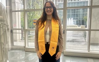 A smiling young woman stands in front of a wall of windows wearing a cardigan and a gold graduation sash reading The Honors College on one end and Unigersity at Albany on the other