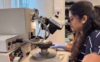 A woman with long dark hair in a blue and white striped blouse looks into a microscope in a laboratory
