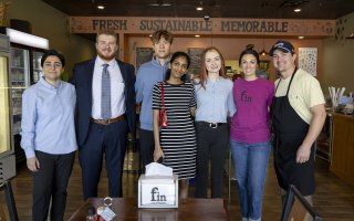 A group of five UAlbany MBA students stand with the owners of fin-your fishmonger inside the store in Guilderland in front of a sign on the wall that says FRESH - SUSTAINABLE - MEMORABLE.