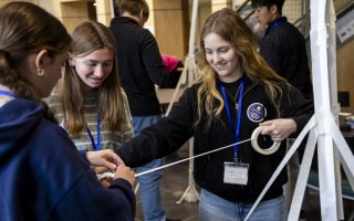 Three smiling teen girls hold onto a spool of masking tape as they attempt to build a freestanding paper tower.