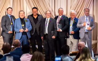 Seven smiling men pose, six of them hold glass awards and wear purple or gold pendants.