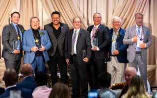 Seven smiling men pose, six of them hold glass awards and wear purple or gold pendants.