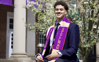 A young man with short curly hair in a suit and purple sash that reads "Spellman Scholar" sits outside on a sunny spring day.