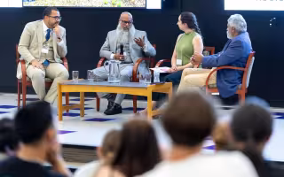 Four people sit around a coffee table set up on a stage. A man with glass and a long beard, wearing a gray suit holds a microphone as he addresses the group. 