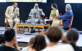 Four people sit around a coffee table set up on a stage. A man with glass and a long beard, wearing a gray suit holds a microphone as he addresses the group. 