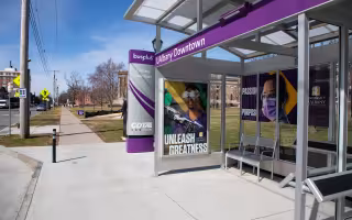 A purple CDTA bus shelter displaying “UAlbany Downtown” and “Unleash Greatness” signage, with metal seating beneath the covered glass structure.
