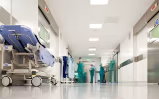 Healthcare workers wearing green scrubs, masks and surgical caps work at the end of a brightly lit hospital hallway.