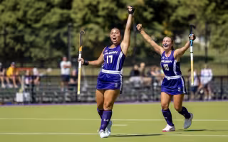 A young woman with dark brown hair in a purple field hockey uniform of the UAlbany Great Danes wearing number 17 lifts her arm in celebration on the turf field.