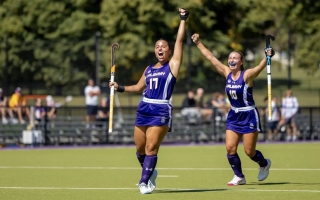A young woman with dark brown hair in a purple field hockey uniform of the UAlbany Great Danes wearing number 17 lifts her arm in celebration on the turf field.