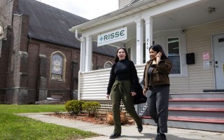 Two women smile as they exit a building with a sign reading: “RISSE Refugee & Immigrant Support Services of Emmaus” hanging from the porch roof. A brick church is visible in the background. 