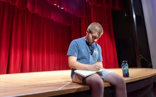 A teen boy sits on a theatre stage in front of a red curtain and writes in a notebook. 
