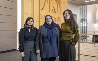 Three women stand inside in front of a large Minera logo from the University at Albany behind them
