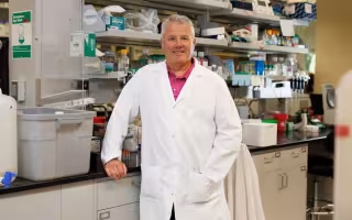 A man wearing a white lab coat and salmon shirt smiles for a portrait in a biology lab.