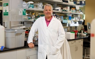 A man wearing a white lab coat and salmon shirt smiles for a portrait in a biology lab.