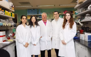 Five people wearing white lab coats smile for a group portrait in a brightly lit biology lab. The group is positioned between two rows of lab benches and shelving containing various containers and biology research equipment.