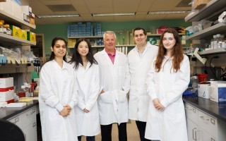 Five people wearing white lab coats smile for a group portrait in a brightly lit biology lab. The group is positioned between two rows of lab benches and shelving containing various containers and biology research equipment.