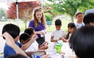 A woman in a purple shirt stands in a group of children making slime in a park under a pop-up tent