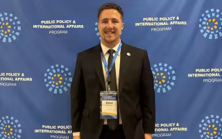 Dylan Repertorio stands in a suit wearing a Public Policy and International Affairs Junior Summer Institute credential.