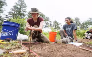 A young woman with a red shirt and cargo pants kneels on the ground next to a young woman in a gray shirt and cargo pants by a bucket and a grid of dirt at an archeological field school in Albany's Pine Bush.