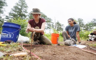 A young woman with a red shirt and cargo pants kneels on the ground next to a young woman in a gray shirt and cargo pants by a bucket and a grid of dirt at an archeological field school in Albany's Pine Bush.