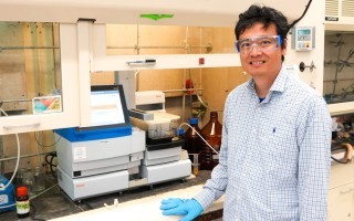 A man wearing a plaid button-down shirt, clear protective goggles and blue latex gloves smiles for a portrait standing in front of a lab bench.