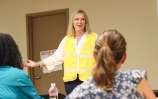 A woman wearing a yellow high-vis vest smiles as she addresses a room full of training participants.