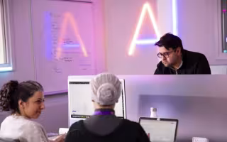 Two student sit facing in each other in front of a computer screen while a professor looks on with the letters "AI" illuminated on a wall behind them.