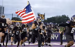 A football player in a black uniform with the word UALBANY on the front carries the American Flag onto the field surrounded by teammates.